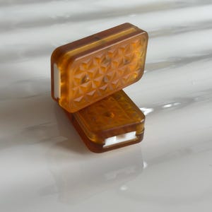 Op de afbeelding: Twee rechthoekige amberkleurige harscomponenten met een geometrisch stervormig patroon op het oppervlak. Elk stuk heeft een doorschijnende bruine afwerking en een contrasterende witte inzet die zichtbaar is langs de zijkant.