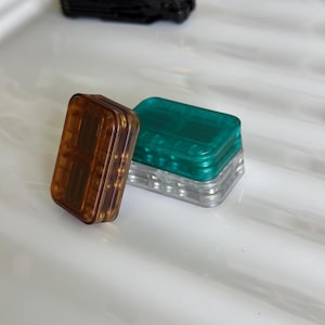 Könnte beinhalten: Zwei rechteckige Fidget-Spielzeuge aus durchscheinendem Kunststoff. Eines ist bernsteinfarben und steht aufrecht, während das andere in Blaugrün und Transparent gehalten ist und flach liegt. Beide zeigen innenliegende mechanische Bauteile.