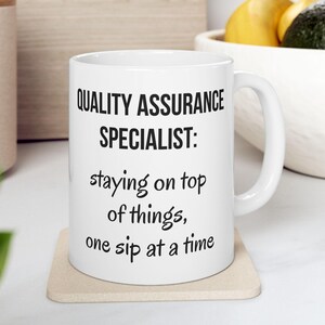 May include: White ceramic coffee mug with black text that reads "QUALITY ASSURANCE SPECIALIST: staying on top of things, one sip at a time".