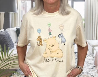 May include: A cream colored t-shirt with a cartoon Winnie the Pooh bear holding a heart. The bear is surrounded by four balloons with the names Emma, Theodore, Samuel, and a fourth balloon with an unreadable name. The text "Mimi Bear" is printed below the bear.