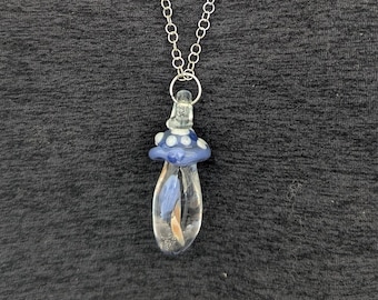 May include: A clear glass mushroom pendant with a blue cap and a silver chain. The mushroom has a blue and white dotted pattern on the cap and a small, dried flower inside. The chain is a silver color.