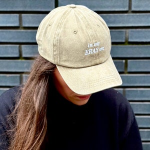 May include: A khaki green baseball cap with the text "in my ERAS era" embroidered in white on the front.