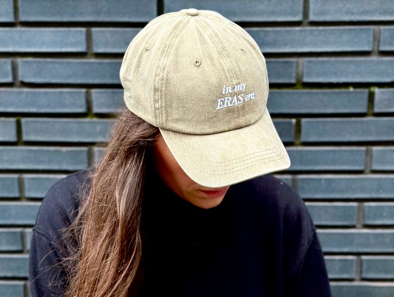 May include: A khaki green baseball cap with the text "in my ERAS era" embroidered in white on the front.
