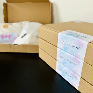 May include: Three brown cardboard boxes stacked on top of each other. Each box has a sticker with a watercolor design and the text "Hi, sweetie, open me!"