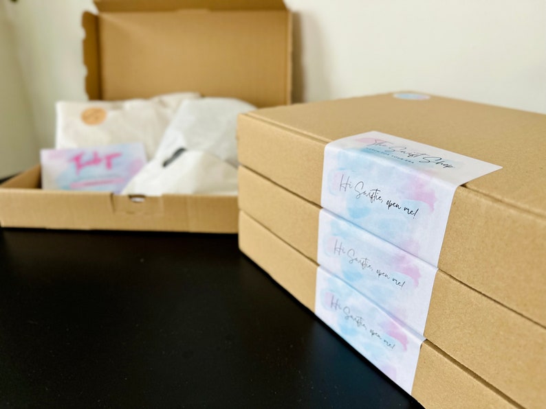 May include: Three brown cardboard boxes stacked on top of each other. Each box has a sticker with a watercolor design and the text "Hi, sweetie, open me!"