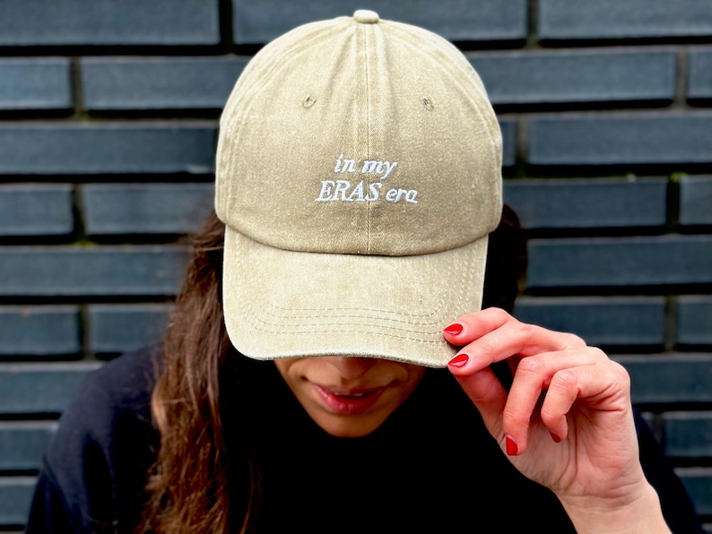 May include: A beige baseball cap with the text "in my ERAS era" embroidered in white on the front.