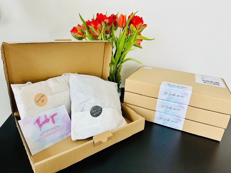 May include: A cardboard box filled with tissue paper and two packages labeled "Pockets Bag" and "Glass Cap". The box is open and a thank you card is visible. There are four stacked cardboard boxes with a label that reads "The Great Shop" and "It's Simple you know!" in a watercolor design.