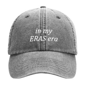 May include: A gray washed denim baseball cap with white embroidered text that reads "in my ERAS era".