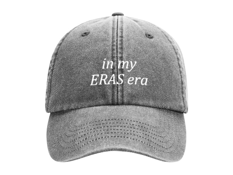 May include: A gray washed denim baseball cap with white embroidered text that reads "in my ERAS era".