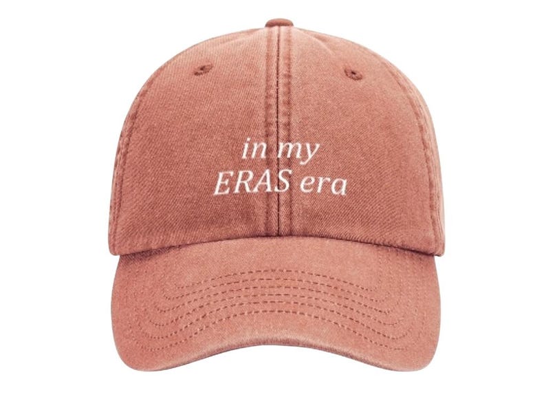 May include: A dusty rose colored baseball cap with white embroidered text that reads "in my ERAS era".