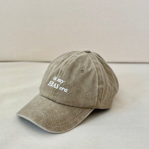 May include: A light brown baseball cap with the text "in my ERAS era" embroidered in white on the front.