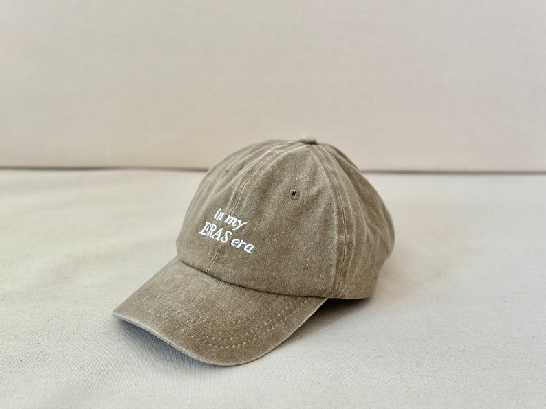 May include: A light brown baseball cap with the text "in my ERAS era" embroidered in white on the front.