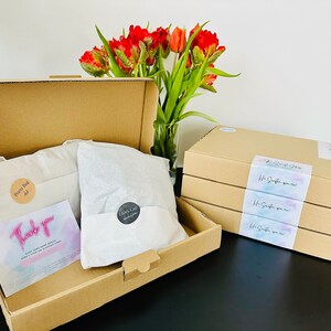 May include: A brown cardboard box with a thank you card and two white tissue paper-wrapped items labeled "Pockets Bag" and "Glass Cap". The box is open and sitting on a table next to a stack of three brown cardboard boxes with a label that says "The Great Shop" and "It's Simple you know!"