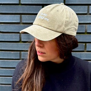 May include: A person wearing a beige baseball cap with the text "In my bras era" embroidered on the front. The person is wearing a black mock neck shirt.