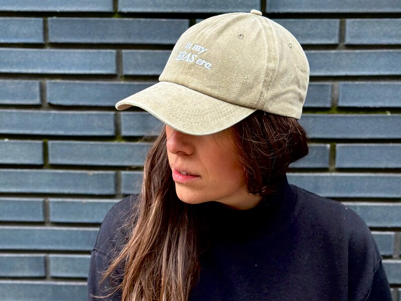 May include: A person wearing a beige baseball cap with the text "In my bras era" embroidered on the front. The person is wearing a black mock neck shirt.