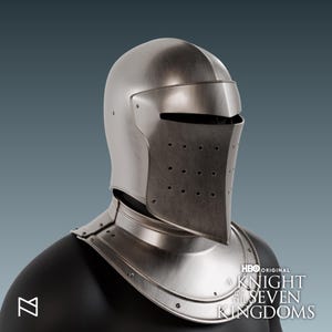 May include: A silver-colored knight's helmet with a visor and neck guard. The helmet has a smooth, metallic finish and small ventilation holes on the visor. The HBO Original logo and "A Knight of the Seven Kingdoms" text are visible.