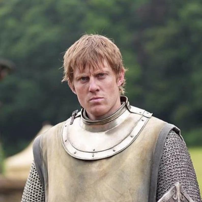 May include: A person with fair skin and reddish-blonde hair wearing medieval armor. The armor includes a metal gorget, chainmail, and a breastplate. The background is blurred, suggesting an outdoor setting.