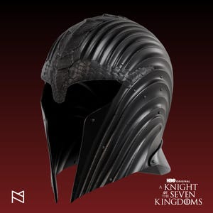 Puede incluir: Casco negro con frente texturizado en forma de serpiente y diseño acanalado. El casco tiene visera y está adornado con remaches. Se ve el logo de HBO Original y el texto "A Knight of the Seven Kingdoms".