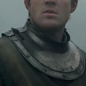 May include: Close-up of a medieval steel gorget, a neck and upper chest armor piece. The gorget is riveted and has a dark, weathered appearance. The armor is worn over a dark garment.