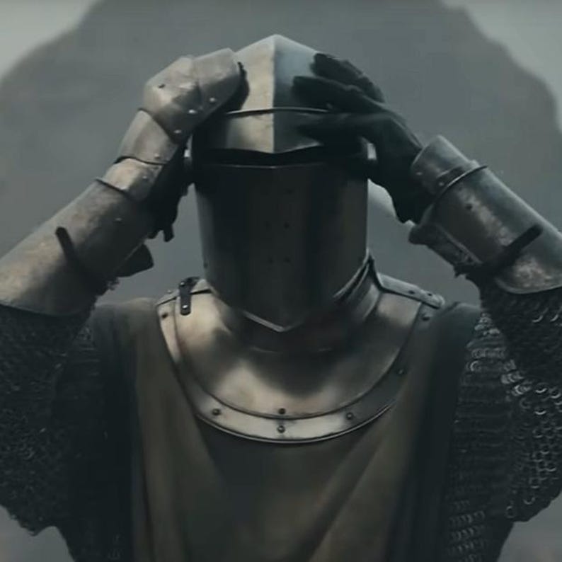 May include: A person wearing a medieval suit of armor, including a helmet, shoulder guards, and chainmail. The armor is a dull silver color, and the person's hands are touching the helmet. The background is a blurred, neutral tone.