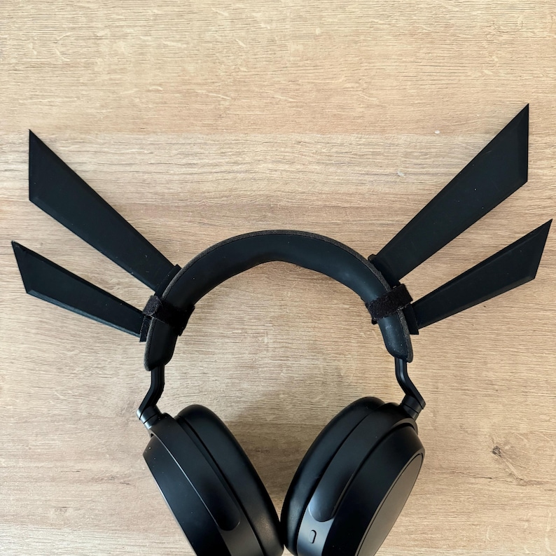 K&ouml;nnte beinhalten: Schwarzer Over-Ear-Kopfh&ouml;rer mit einem individuellen Aufsatz mit drei schwarzen, eckigen Klingen. Der Kopfh&ouml;rer hat ein gepolstertes Kopfband und Ohrmuscheln. Der Aufsatz wird mit Klettb&auml;ndern befestigt. Der Hintergrund ist eine hellbraune Holzoberfl&auml;che.