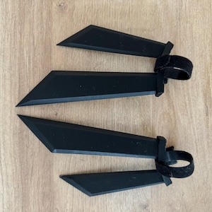K&ouml;nnte beinhalten: Vier schwarze, kantige Objekte mit klingenartiger Form, jeweils verbunden mit einem schwarzen Riemen mit Klettverschluss. Die Objekte sind auf einer hellbraunen Holzoberfl&auml;che angeordnet.