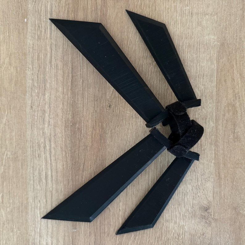 K&ouml;nnte beinhalten: Vier schwarze, abgewinkelte Klingen mit einem zentralen Klettverschluss, angeordnet in einem f&auml;cherartigen Muster. Die Klingen haben ein scharfes, spitzes Ende und bestehen aus einem mattschwarzen Material. Das Objekt liegt auf einer hellbraunen Holzoberfl&auml;che.