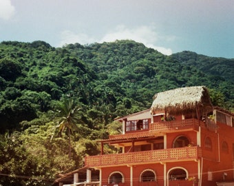 May include: A multi-story, reddish-orange building with a thatched roof sits on a beach, backed by a lush green hillside. A bird soars in the sky above. The building has balconies and arched windows, with a small boat on the sand.