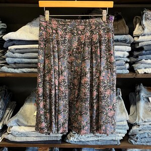 May include: A vintage floral print skirt in shades of navy, pink, and green, hanging on a wooden hanger. The skirt features a pleated design and a delicate floral pattern. The background shows shelves of denim jeans.