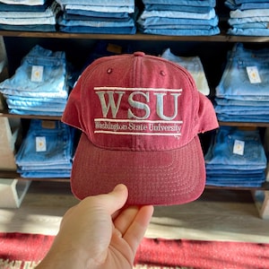 May include: A maroon baseball cap with the letters "WSU" embroidered in gray, along with "Washington State University." The cap is held in front of a shelf stacked with blue jeans.