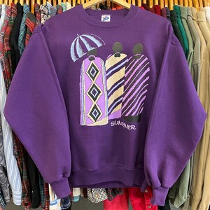 May include: A purple sweatshirt with a graphic print featuring three figures under an umbrella. The figures are wearing patterned robes in shades of purple, yellow, and black. The word "SUNRIVER" is printed below the figures.