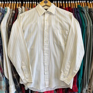 May include: A button-down shirt with vertical stripes in light blue and beige. The long-sleeved shirt has a chest pocket and is hanging on a wooden hanger. The shirt is part of a collection of clothing on a rack.