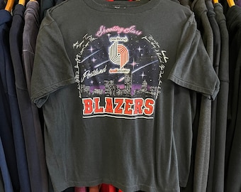 Puede incluir: Camiseta gris oscuro con un diseño gráfico de los Portland Trail Blazers. El diseño incluye las palabras "Shooting Stars" y "Blazers" con un paisaje urbano e imágenes de baloncesto. La camiseta es de manga corta.