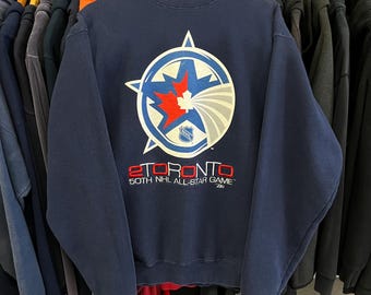 May include: Navy blue sweatshirt featuring a large circular graphic with a starburst design in white, red, and blue. The graphic includes the text "TORONTO" and "50TH NHL ALL-STAR GAME". A vintage sports apparel item.