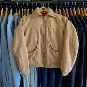 May include: A tan wool-blend jacket featuring a pointed collar, a full-length silver metal front zipper, and two curved front pockets. The garment includes ribbed knit cuffs and a ribbed hem, with a vibrant red interior lining visible at the collar.