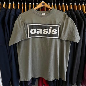 May include: Olive green t-shirt with the word "oasis" in white block letters inside a black rectangle. The shirt is hanging on a wooden hanger among other clothing items. The t-shirt is a vintage style.