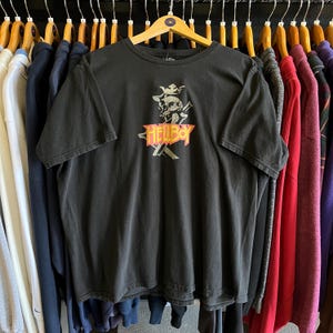 May include: Black t-shirt with a graphic featuring a skull wearing a crown, crossed bones, and the word "HELLBOY" in yellow and orange. The shirt is hanging on a rack with other clothing.