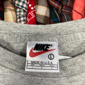 Pu&ograve; includere: Primo piano di un'etichetta di una maglietta Nike grigia. L'etichetta presenta il logo Nike in rosso e bianco, con la scritta "MADE IN U.S.A." e informazioni sulla taglia. La maglietta &egrave; taglia L.