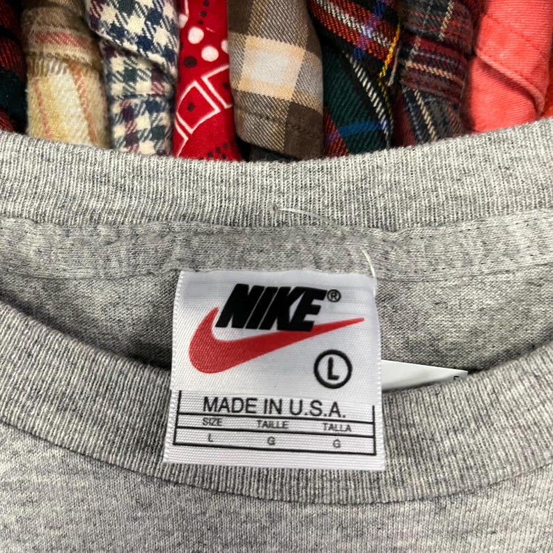 Pu&ograve; includere: Primo piano di un'etichetta di una maglietta Nike grigia. L'etichetta presenta il logo Nike in rosso e bianco, con la scritta "MADE IN U.S.A." e informazioni sulla taglia. La maglietta &egrave; taglia L.