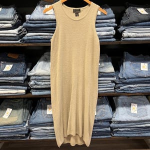 May include: A beige sleeveless dress hangs on a wooden hanger. The dress is made of a textured fabric and has a scoop neckline. The dress is displayed in a vintage clothing store, surrounded by stacks of blue jeans.