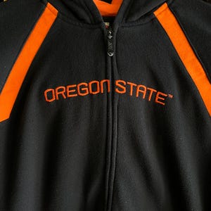 Peut inclure: Sweat &agrave; capuche zipp&eacute; noir avec des accents orange et le texte brod&eacute; "OREGON STATE". Le sweat-shirt a une fermeture &eacute;clair compl&egrave;te et une capuche. Les accents orange sont sur les &eacute;paules.