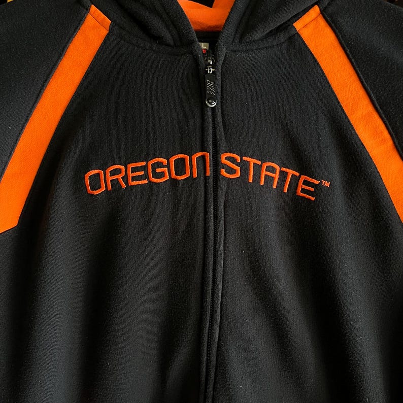 Peut inclure: Sweat &agrave; capuche zipp&eacute; noir avec des accents orange et le texte brod&eacute; "OREGON STATE". Le sweat-shirt a une fermeture &eacute;clair compl&egrave;te et une capuche. Les accents orange sont sur les &eacute;paules.