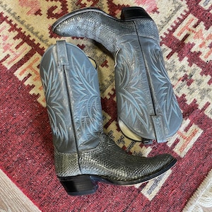 May include: A pair of gray cowboy boots with intricate blue stitching on the shafts. The boots have a textured pattern on the foot and a classic pointed toe. The boots are displayed on a red and cream patterned rug.