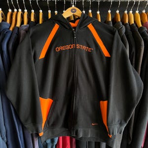 Peut inclure: Sweat &agrave; capuche zipp&eacute; noir avec des d&eacute;tails orange et le texte "OREGON STATE" sur la poitrine. Il poss&egrave;de une capuche, des poches avant et un petit logo Nike. Le sweat est suspendu &agrave; un cintre en bois.