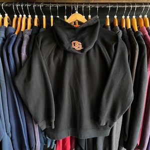 Peut inclure: Sweat &agrave; capuche noir avec les lettres "CS" en orange sur la capuche. Le sweat-shirt est suspendu &agrave; un portant avec d'autres v&ecirc;tements. Le sweat-shirt a un cordon de serrage et une poche kangourou.