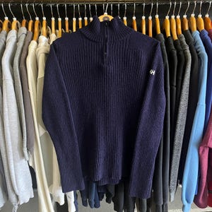 May include: A navy blue ribbed knit sweater featuring a mock neck collar with a short front zipper closure. The garment has long sleeves with wide cuffs and a small white HH logo embroidered on the upper left sleeve, hanging on a wooden hanger.
