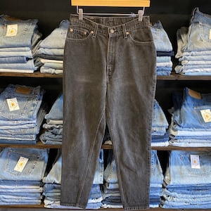 May include: Dark gray denim jeans hanging on a wooden hanger. The jeans have a classic five-pocket design and a button and zipper closure. The background features shelves stacked with folded denim jeans and leather belts.