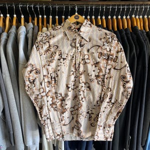 May include: A long-sleeved button-down shirt featuring a cream base with vertical pinstripes and a dark brown floral pattern. The shirt has a standard collar, a front button placket, and buttoned cuffs, displayed on a wooden hanger.