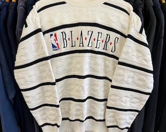 Puede incluir: Suéter de punto color crema con rayas horizontales negras y la palabra "BLAZERS" en el pecho. El suéter presenta un diseño de punto de cable y el logotipo de la NBA. Un suéter de cuello redondo y manga larga de estilo vintage.