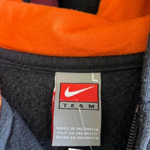 Peut inclure: Gros plan d'un sweat &agrave; capuche noir avec une doublure int&eacute;rieure orange. L'&eacute;tiquette Nike Team est visible, avec le logo Nike et le texte "TEAM". Le sweat-shirt a une fermeture &eacute;clair et une &eacute;tiquette de taille "L".